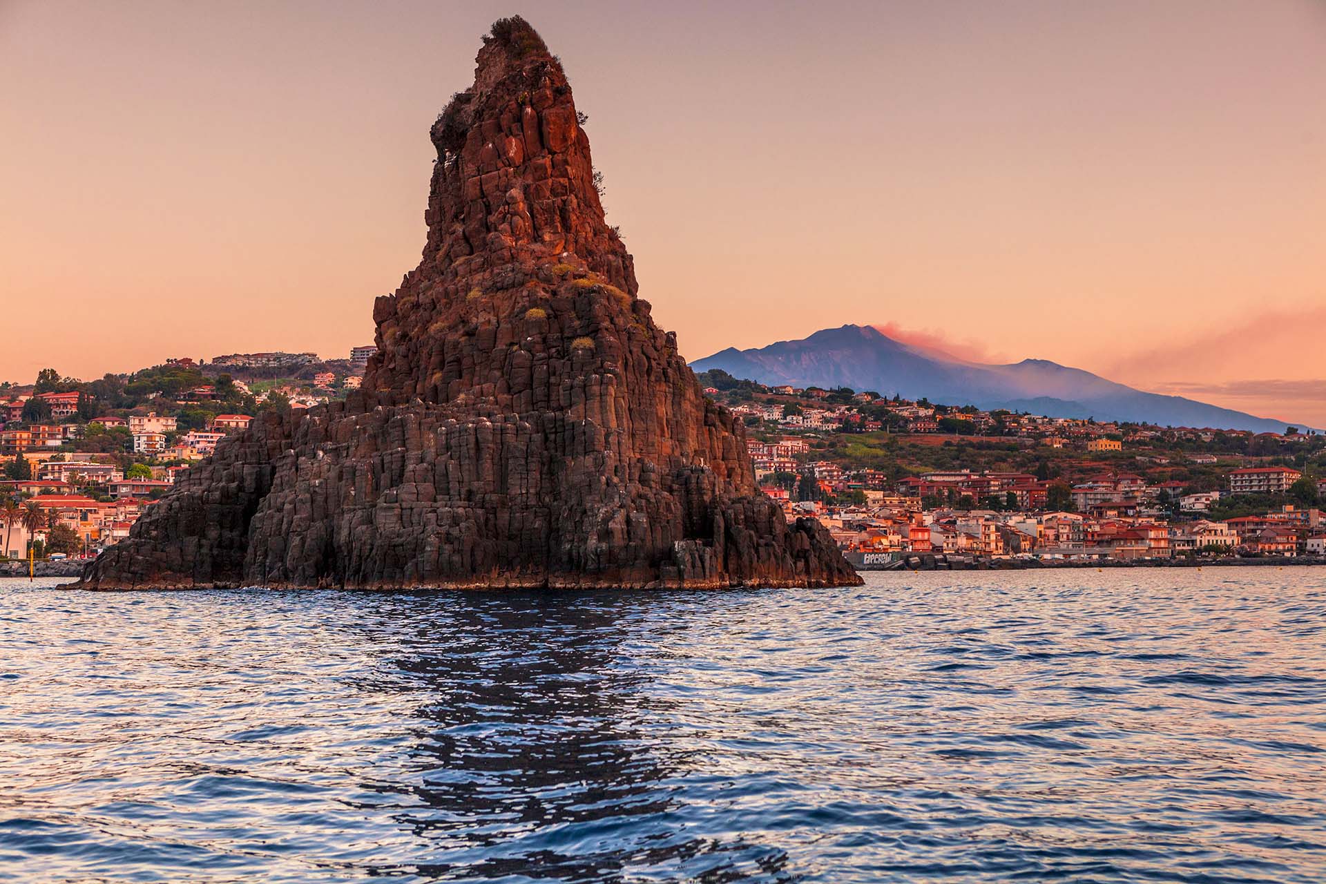 Un paesaggio suggestivo che unisce la cima innevata del vulcano Etna in eruzione e i faraglioni dei Ciclopi che emergono dal mare di Aci Trezza.