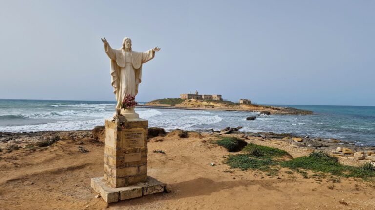 Isola delle Correnti at sunset, with its solitary lighthouse standing out against the colorful sky at the point where the Ionian and Mediterranean seas meet.